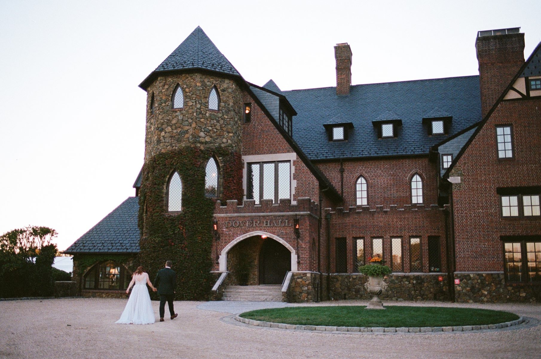 A woman in a white wedding dress and a man in a black tuxedo are walking together and holding hands they are walking toward their wedding venue which is a large mansion made of brick 