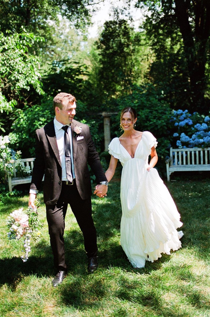 A woman in a white wedding dress is smiling and holding hands with her partner who is looking at her who is wearing a black tuxedo and a black tie and holding a bouquet of flowers 