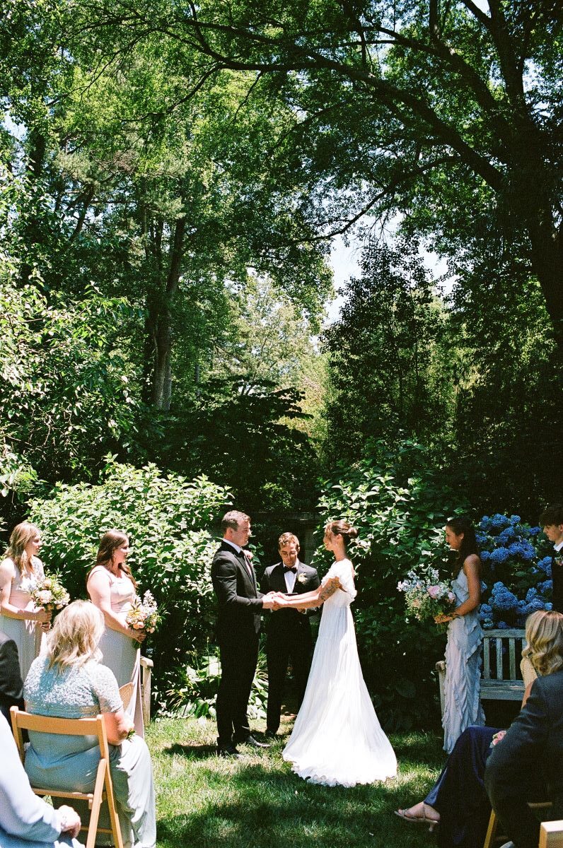 A woman in a white wedding dress is getting married and holding hands with her partner who is wearing a black tuxedo and their guests and wedding party are at their ceremony with the officiant 