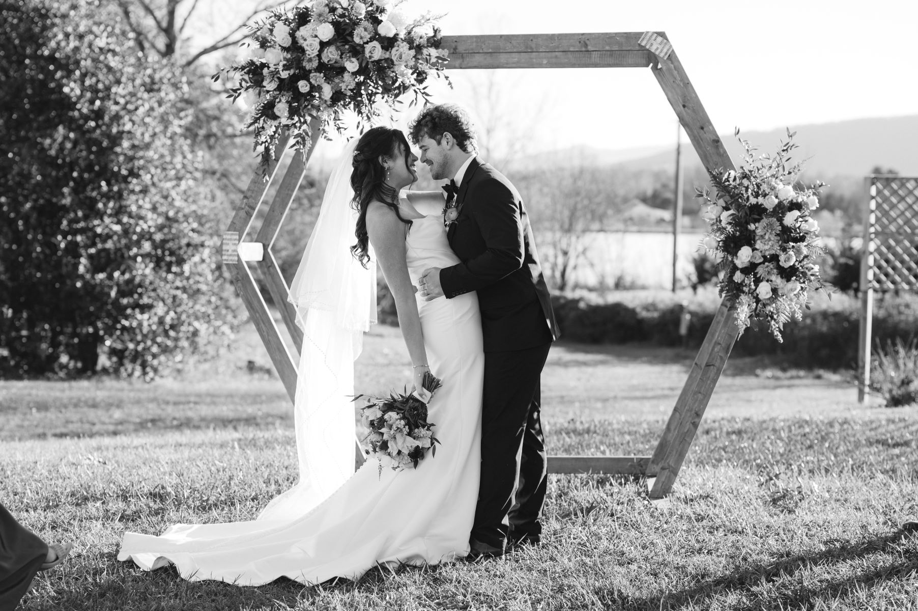 A couple at their wedding ceremony in front of a wooden hexagon shaped arch decorated with flowers the groom is wewaring a black tuxedo and dipping the bride backwards who is wearing a white veil and a white dress and holding a bouquet of flowers in one hand both are smiling at each other 