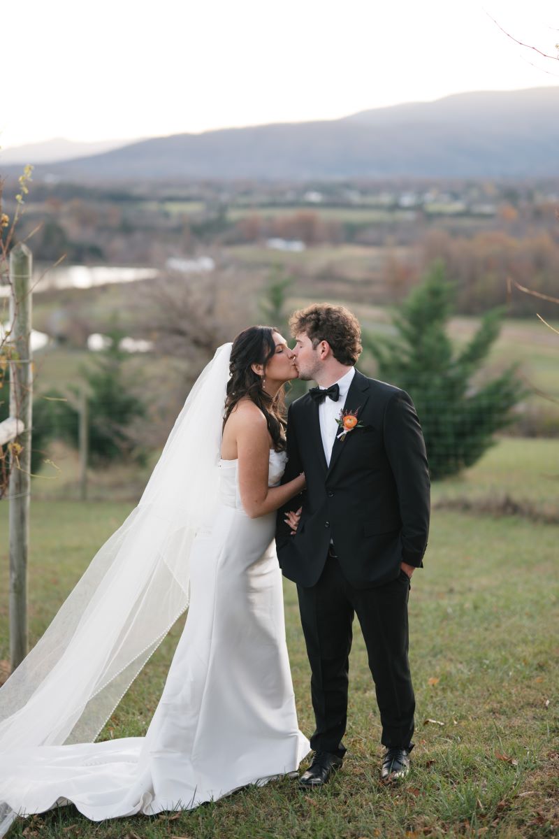 A couple kissing in a vineyard the woman is wearing a white dress and white veil and the man is wearing a black tuxedo and black bowtie