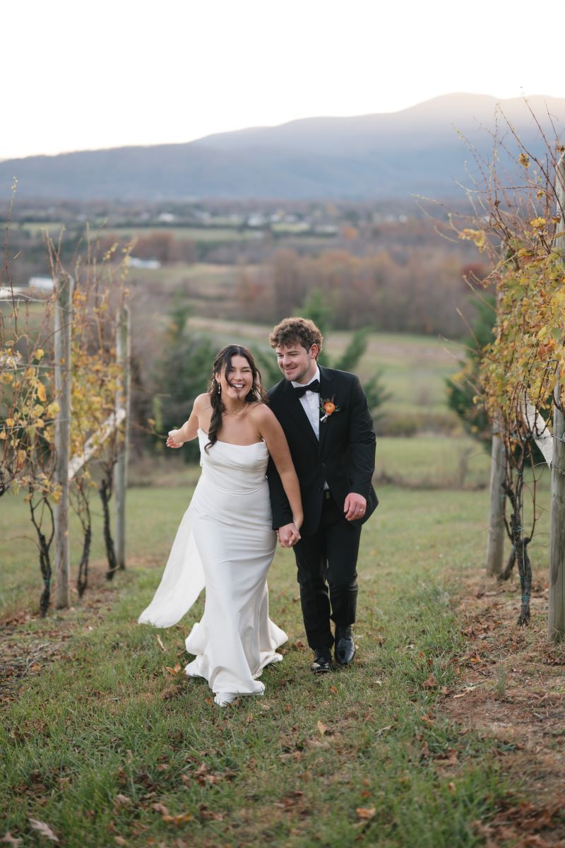 A couple walking through a vineyard the woman is wearing a white wedding dress and white veil and her partner is wearing a black tuxedo and they are holding hands 