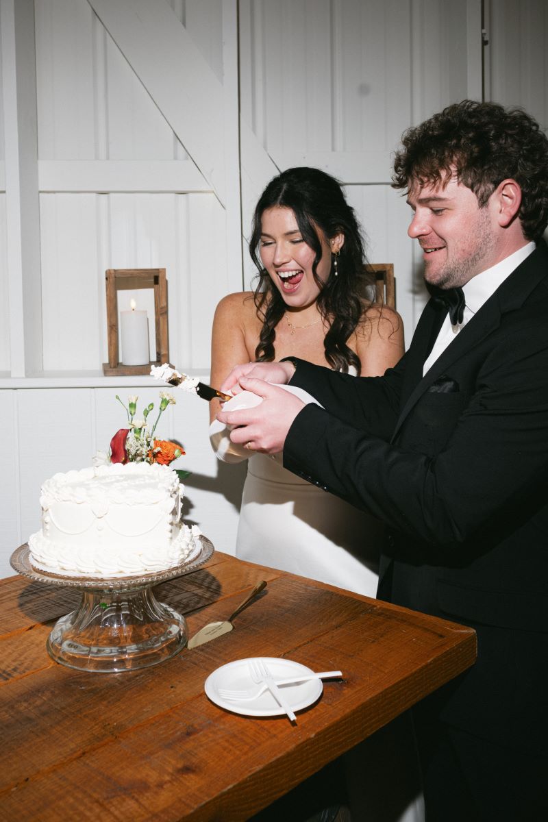 A couple cutting their wedding cake and laughing the man is wearing a black tuxedo and black bowtie and woman is wearing a white wedding dress the cake is white and on a glass platter on a wooden table 