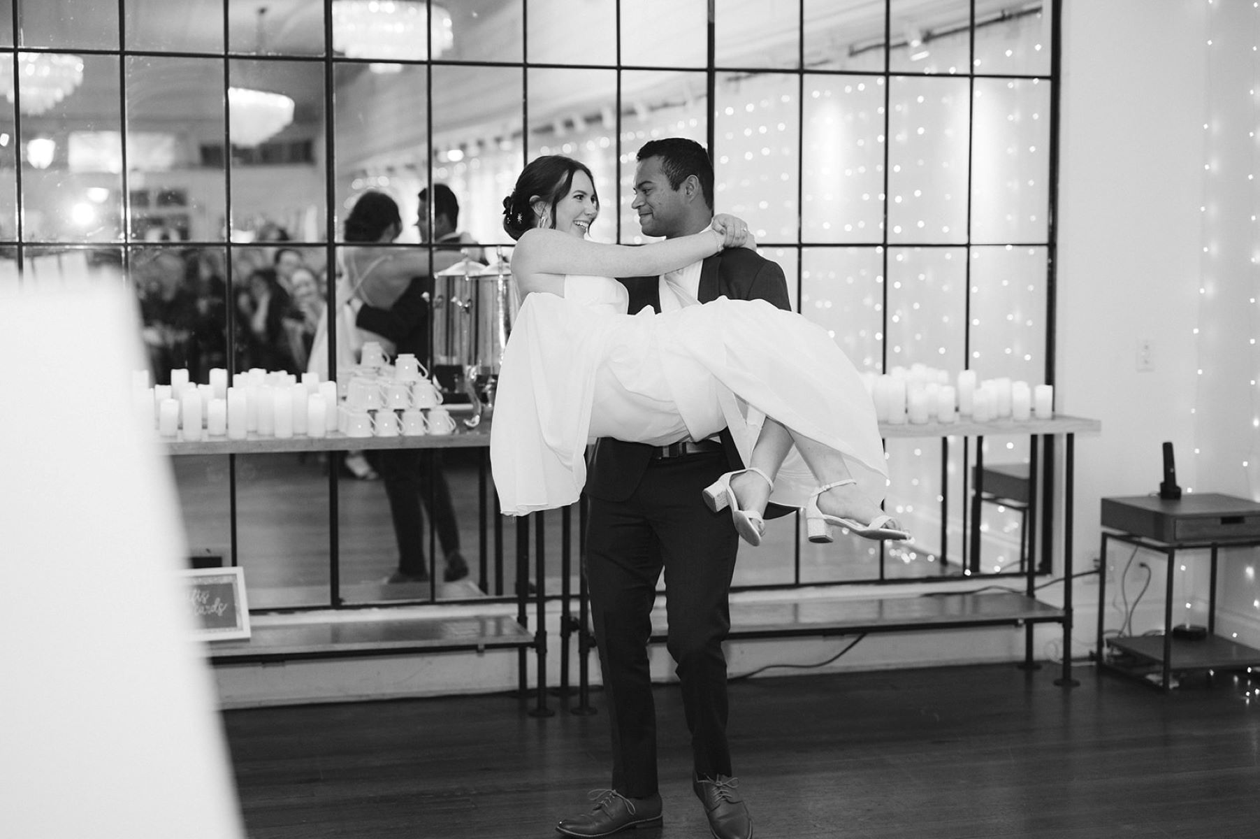 A couple at their wedding reception the husband is wearing a black tuxedo and holding his wife in his arms who is wearing a white dress and white heels and they are both looking at each other and smiling behind them is a table with candles and wall with a glass mirror reflecting their guests watching them 