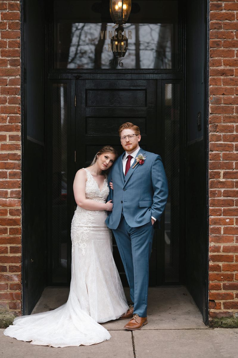 A couple standing in front of an entrance that is made of brick with a black door the woman is wearing a white wedding dress and holding her partner's arm and leaning her head on his shoulder her partner has on a blue tuxedo and a red tie and is wearing glasses he is standing with his hands in his pockets 