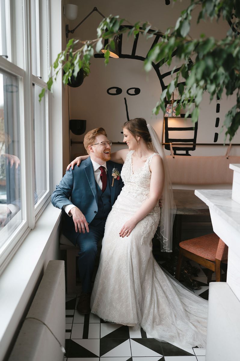 A couple sitting on a chair the woman is wearing a white wedding dress and veil and laughing with her partner who is wearing a blue tuxedo and red tie and has his arm propped against a window sill 