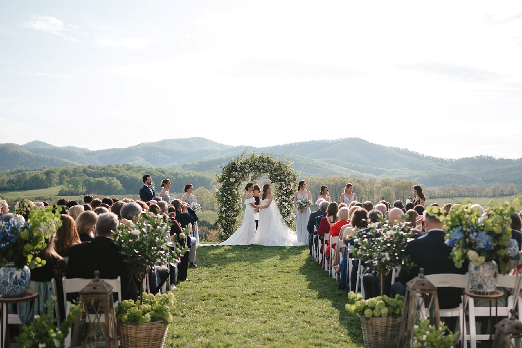 A couple getting married behind them is a floral arch and mountains and in front of them are their wedding guests sitting in white chairs both of the women are wearing white wedding dresses and holding hands and their wedding parties are both standing beside them