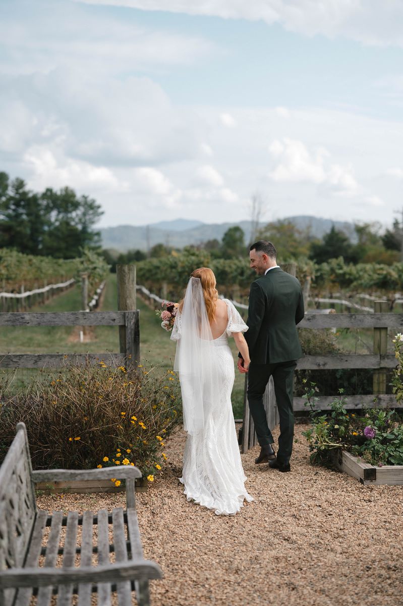 A couple walking towards a vineyard the woman is wearing a white wedding dress and a white veil and holding a bouquet of flowers in one hand with her other hand she is holding her partner's hand who is wearing a green tux 