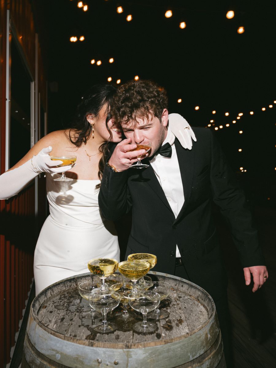 A couple celebrating at their wedding reception the woman is holding her drink in her hand and hugging her partner and smiling she is wearing white gloves and a white dress her partner is taking a sip of his drink and he is wearing a black tuxedo and black bowtie and in front of them is a wooden barrel with additional drinks 