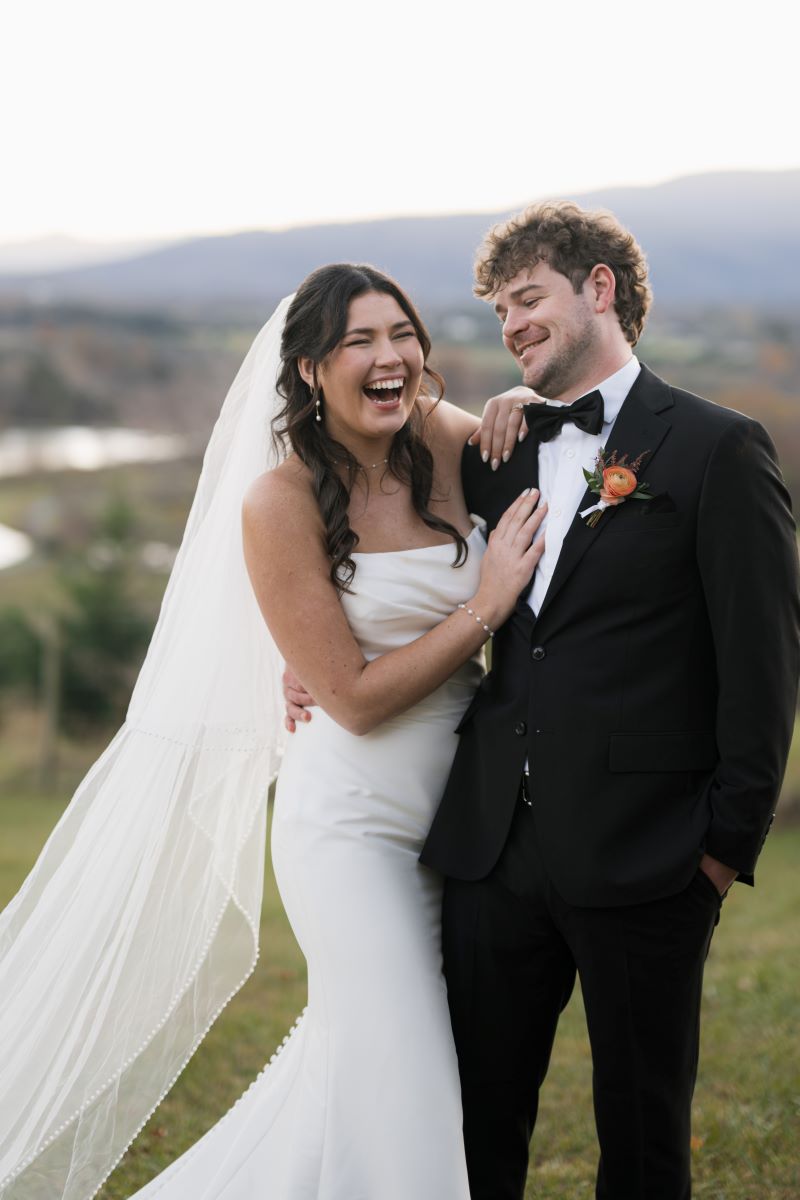 A couple in a vineyard laughing with each other the woman is wearing a white wedding dress and white veil and has her hand on her partner's chest who is wearing a black tuxedo and black bowtie 