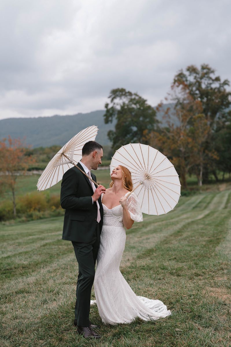 A couple in a field of grass holding white umbrellas and they are both smiling at each other the woman is wearing a white wedding dress the man is wearing a green tuxedo 