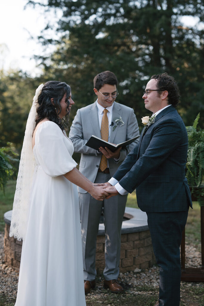 The couple holds hands during their wedding ceremony at Camp Holiday Trails, standing by a stone fire circle and surrounded by greenery as the officiant smiles.