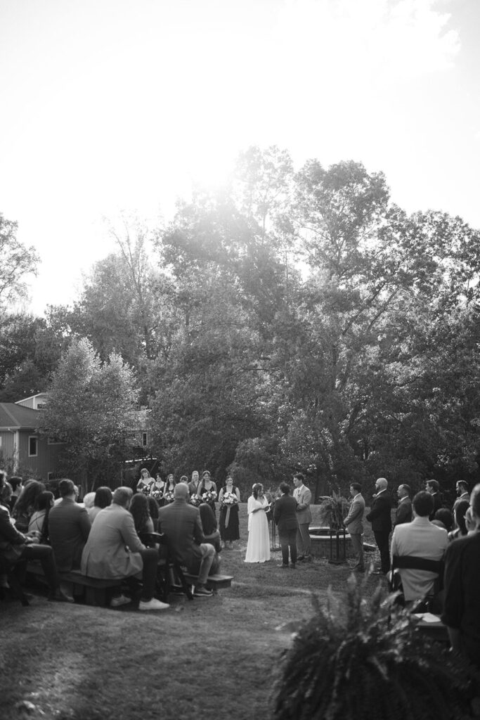 The outdoor wedding ceremony at Camp Holiday Trails, with guests seated on wooden benches under the tall trees.