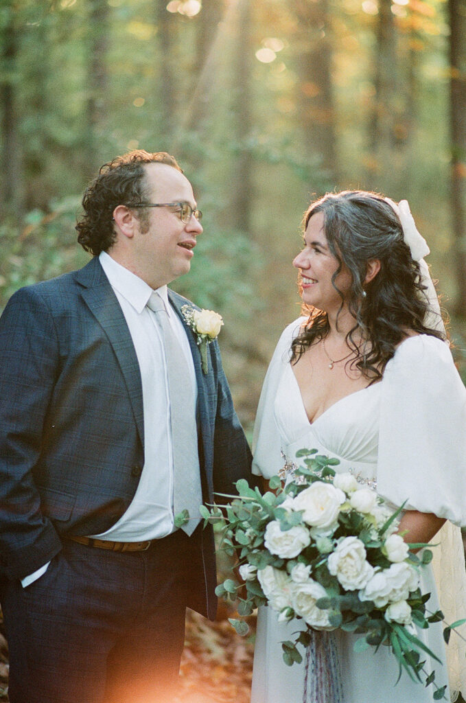 The groom in a suit and tie smiles at his bride, who wears a flowing white gown with puffed sleeves and holds a bouquet of white roses.