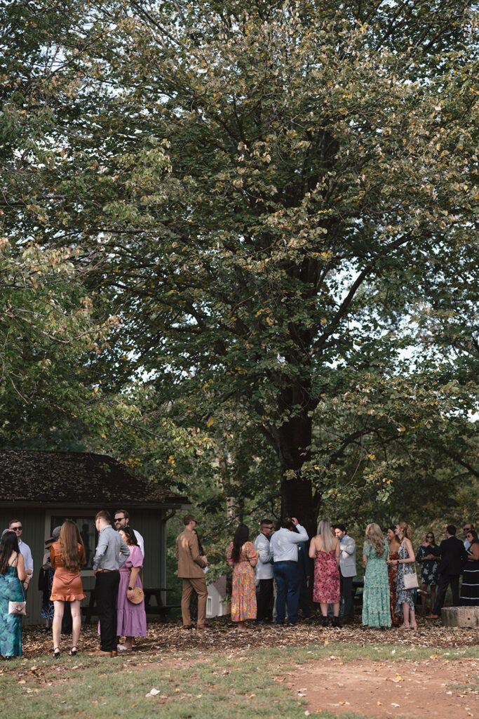 Guests dressed in colorful and semi-formal attire chat under the shade of a large tree near a building at Camp Holiday Trails. 