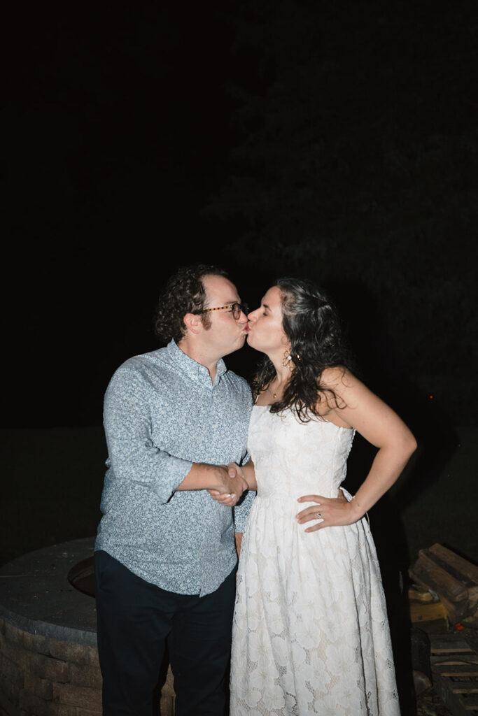 The couple shares a kiss at night, standing beside a glowing campfire.