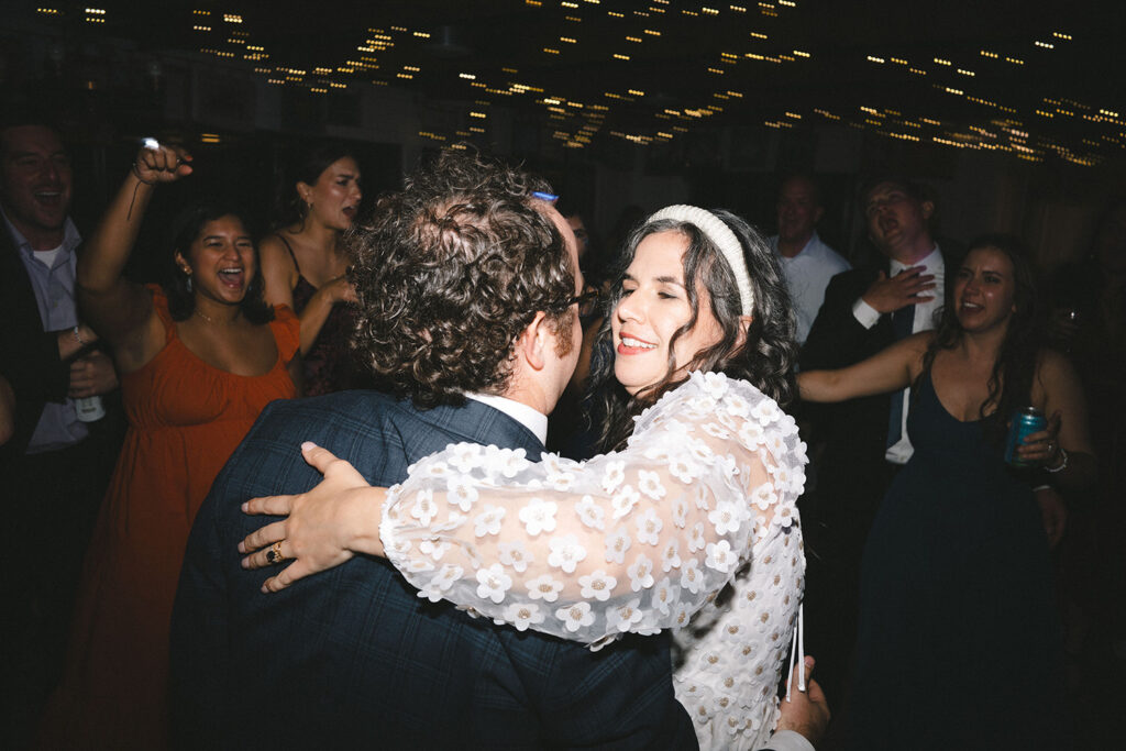 A joyous moment of the couple dancing closely, surrounded by cheering friends and family, with golden string lights.