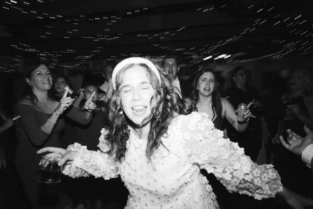 A recently-married person, wearing a floral-textured dress with a headband, energetically dancing with guests under string lights at the reception at Camp Holiday Trails.