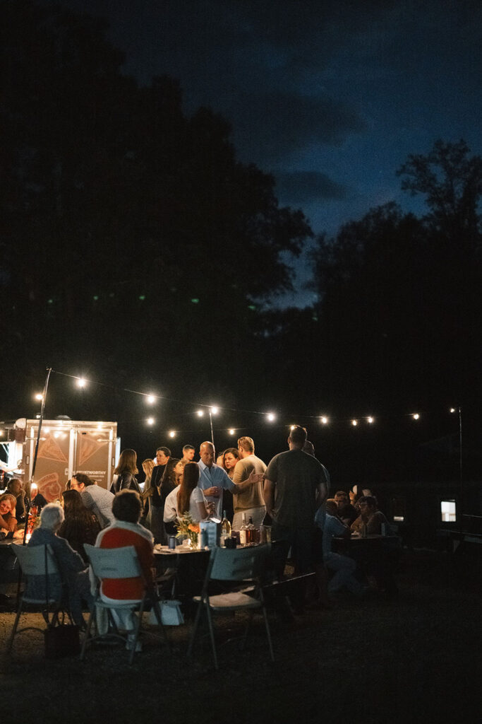 A cozy evening gathering at Camp Holiday Trails with guests mingling under string lights, enjoying an outdoor dining setup surrounded by the night sky.