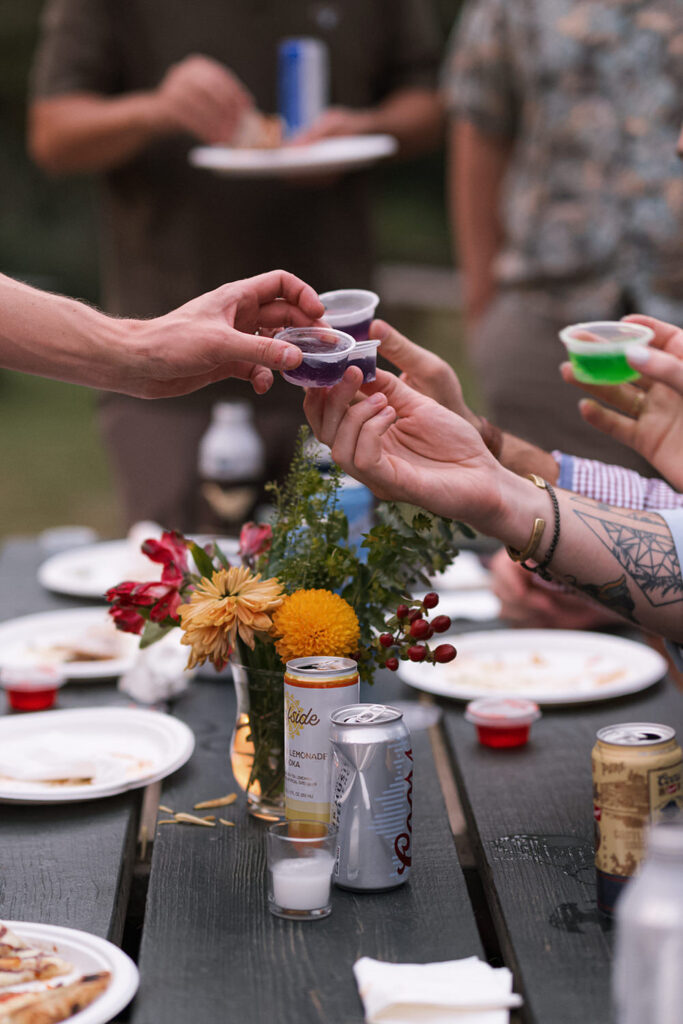 Close-up of hands raising colorful jello shots in celebration, with a picnic table.