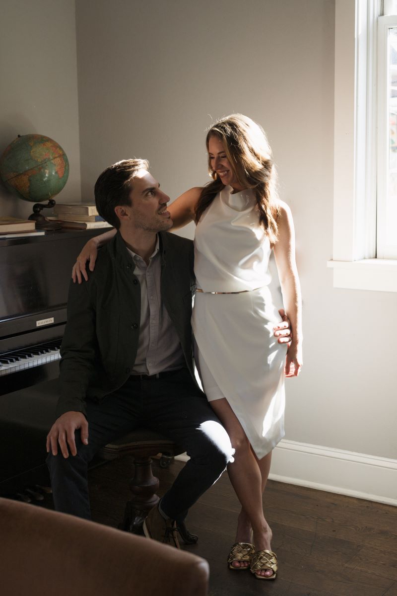 A couple in a room next to a piano the woman is wearing a white dress and standing up the man is sitting down on the pian bench they are both looking at each other and smiling 