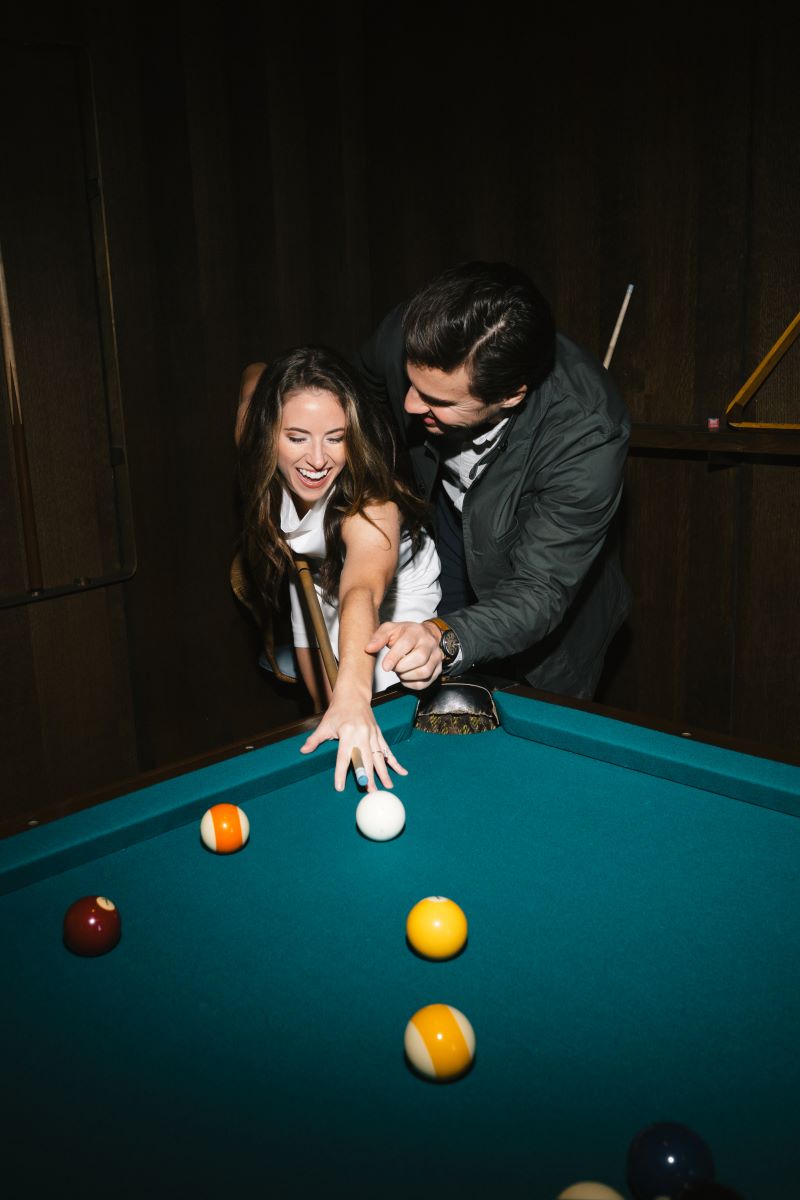 A couple playing pool together the woman is wearing a white dress and holding the pool stick and her partner is guiding her 