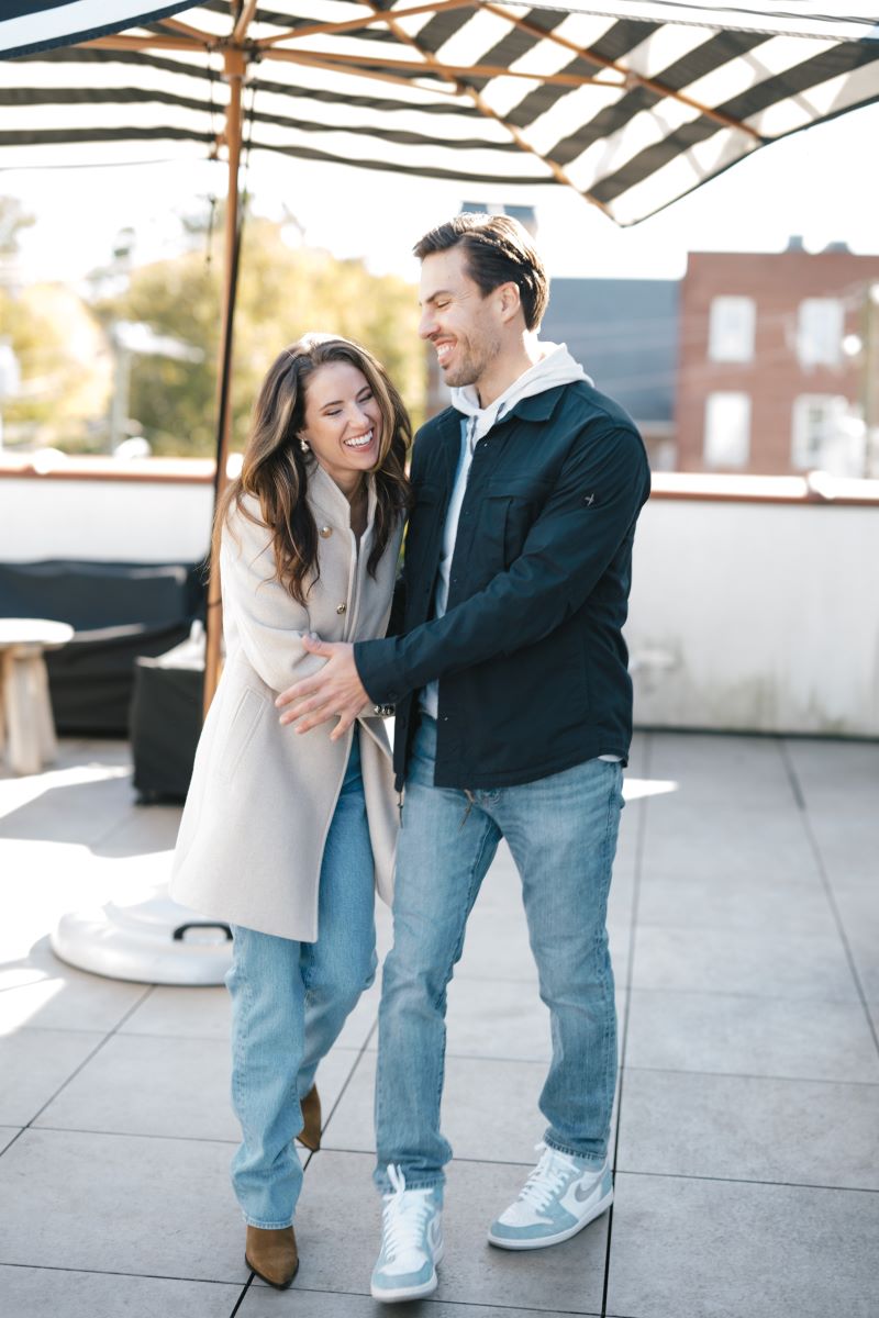 A couple on a terrace laughing together underneath a black and white striped umbrella the woman is wearing a tan jacket and jeans and her partner is wearing a hoodie and blue jacket and jeans and they are hugging each other 