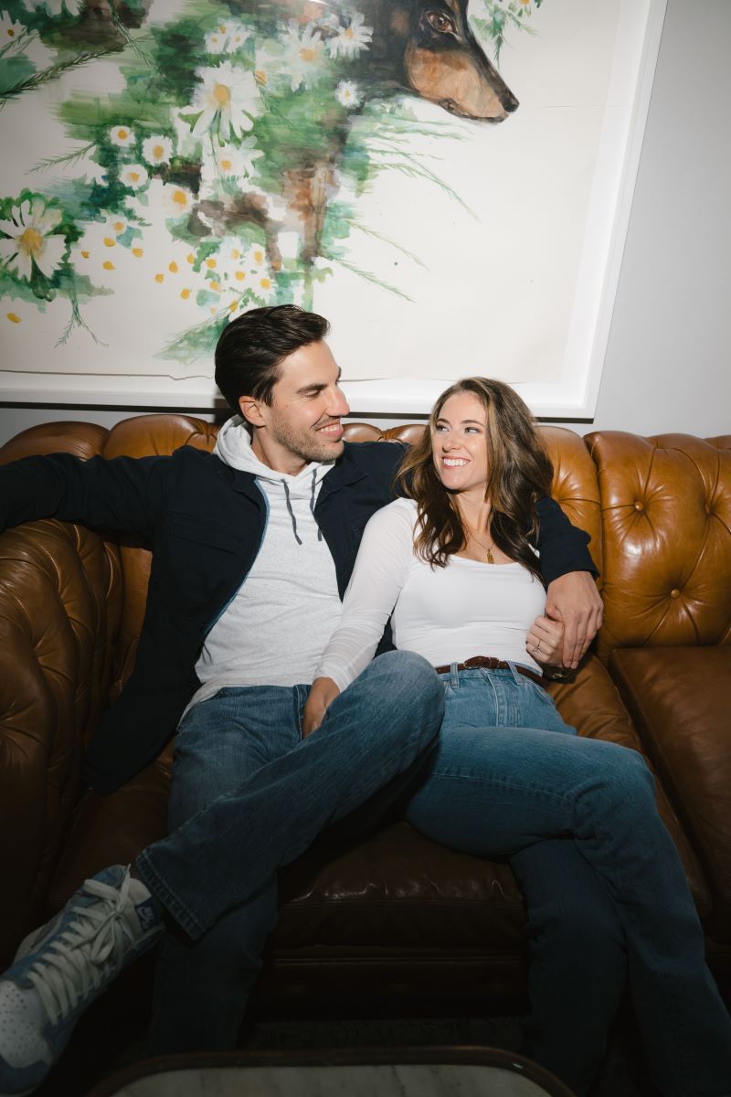 A couple sitting on a borwn leather couch together both are smiling and the woman is grabbing her partner's leg her partner has his arm around her shoulder they are both wearing blue jeans and the woman is wearing a white top the man is wearing a white hoodie and black jacket 