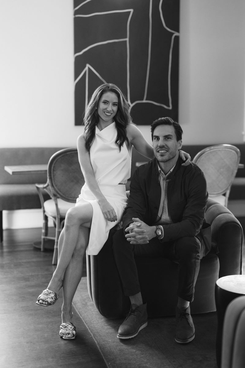 A couple sitting together in an arm chair the woman is wearing a white dress and sitting on the arm rest her partner is sitting next to her 