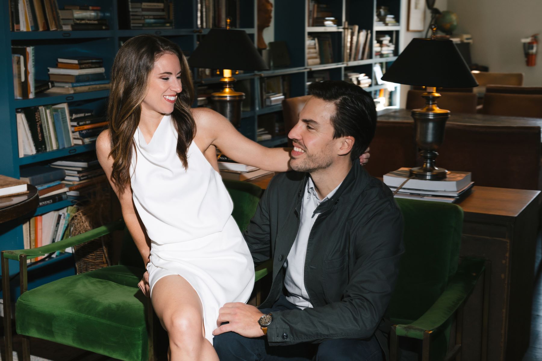 A couple sitting together in a library the woman is wearing a white dress and the man is wearing a black jacket and white shirt the man is sitting on a green arm chair and the woman is sitting perched on the arm rest the couple is smiling at each other