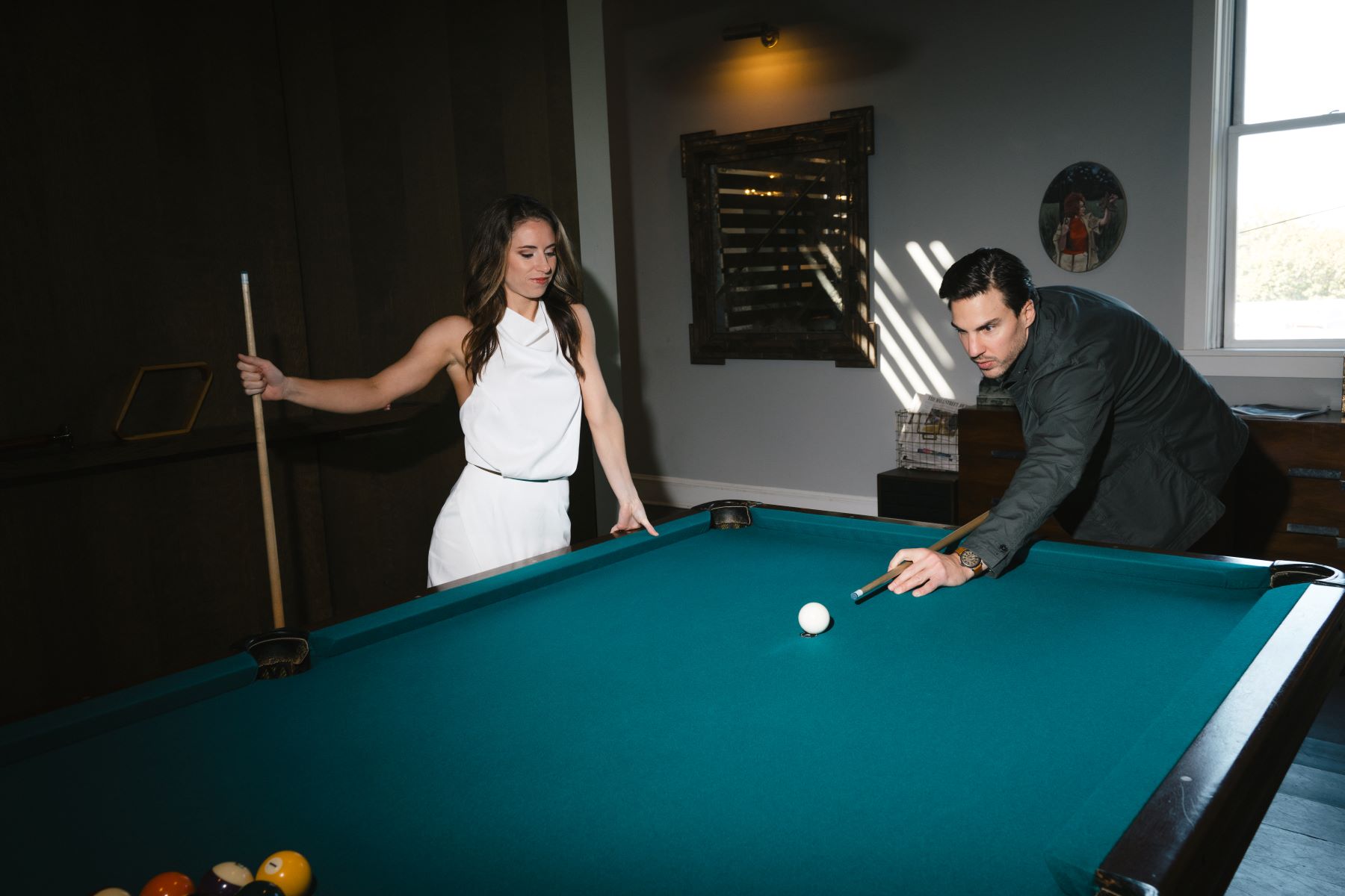 A couple playing pool together they are both holding pool sticks the woman is leaning against the pool table and watching her partner hit the white pool ball