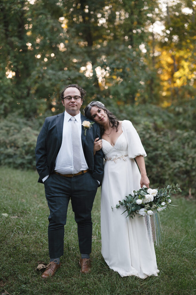 A couple portrait in natural light at Camp Holiday Trails, with the groom in a navy plaid suit and the bride in a simple white gown with puff sleeves, holding a bouquet of white roses and greenery.