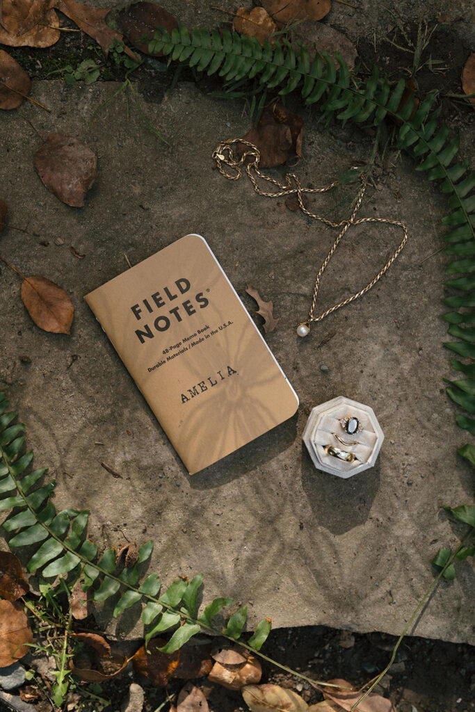 A close-up shot of wedding details at Camp Holiday Trails, featuring a Field Notes journal labeled "Amelia," a pearl necklace, and a jewelry box with rings, surrounded by fern leaves and scattered foliage on a stone background.