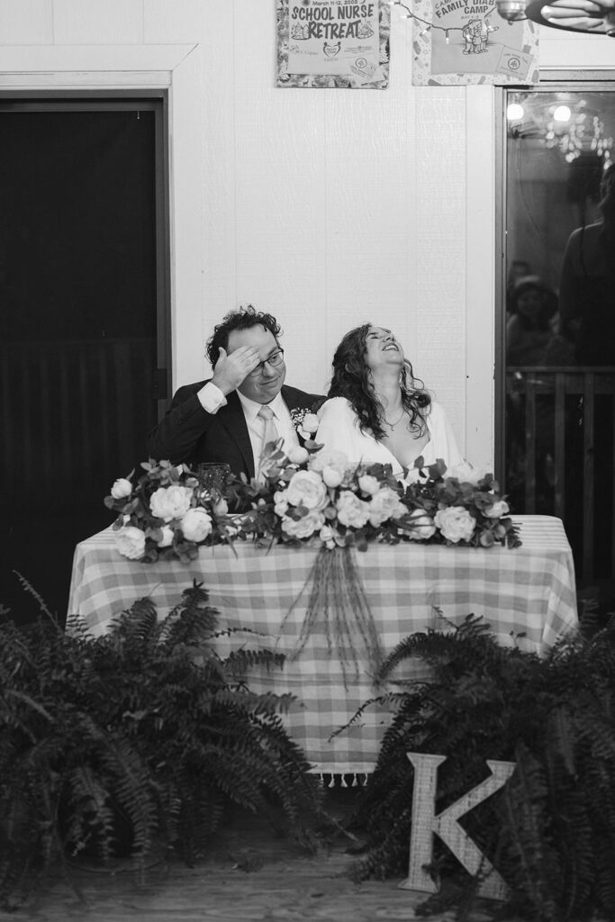 A couple seated at their wedding table at Camp Holiday Trails, laughing together. The table is decorated with a gingham tablecloth, floral arrangements, and ferns.