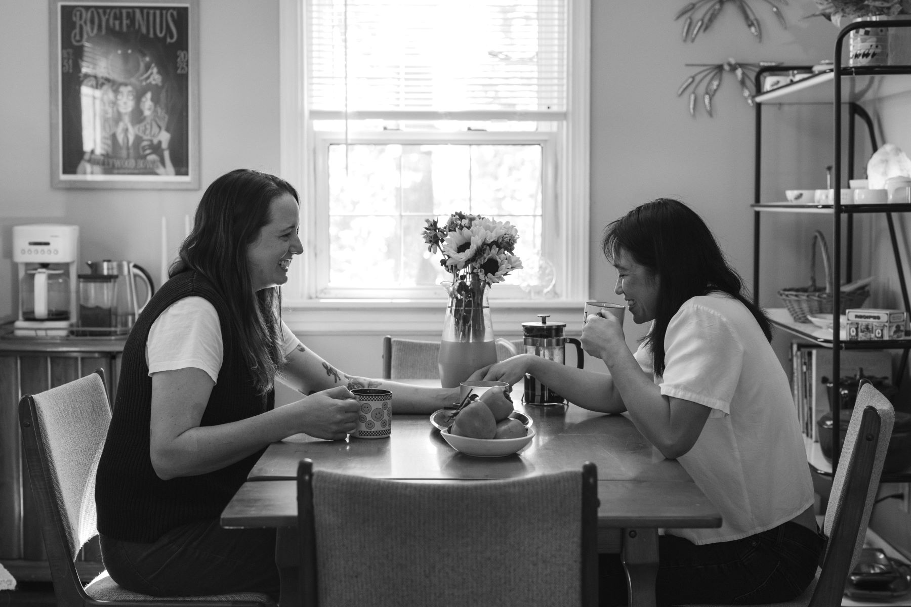 A couple sitting at a kitchen table drinking coffee, laughing, and holding hands. 