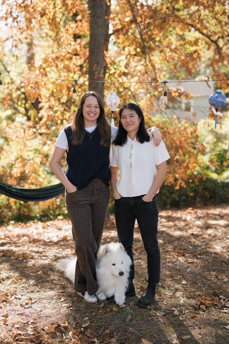 A couple standing in their backyard one person has their arm around the other and their dog is laying on the ground between them 