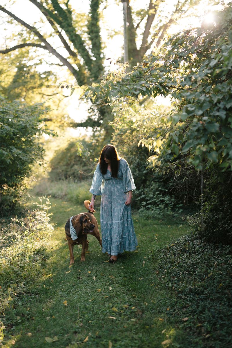 A woman in a long flowy blue dress walking through a field with her dog and the field is surrounded by trees 