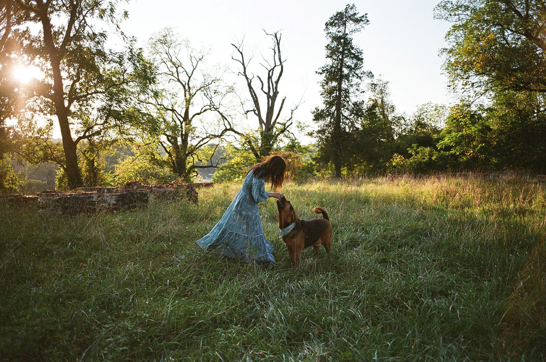 A woman in a field surrounded by trees who is wearing a long flowy blue dress and leaning down to pet her dog