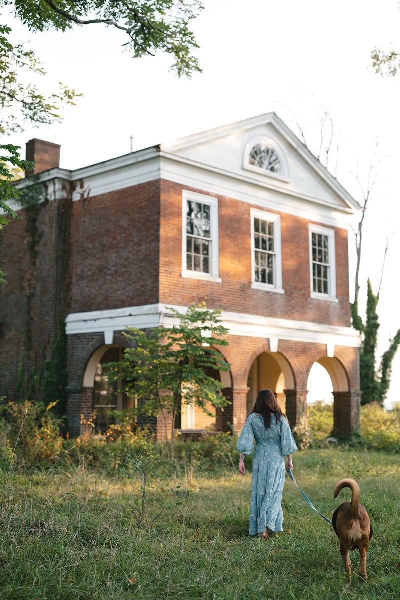 A woman in a long flowy blue dress walking with her dog who is on a leash through the grass towards a brick hourse 