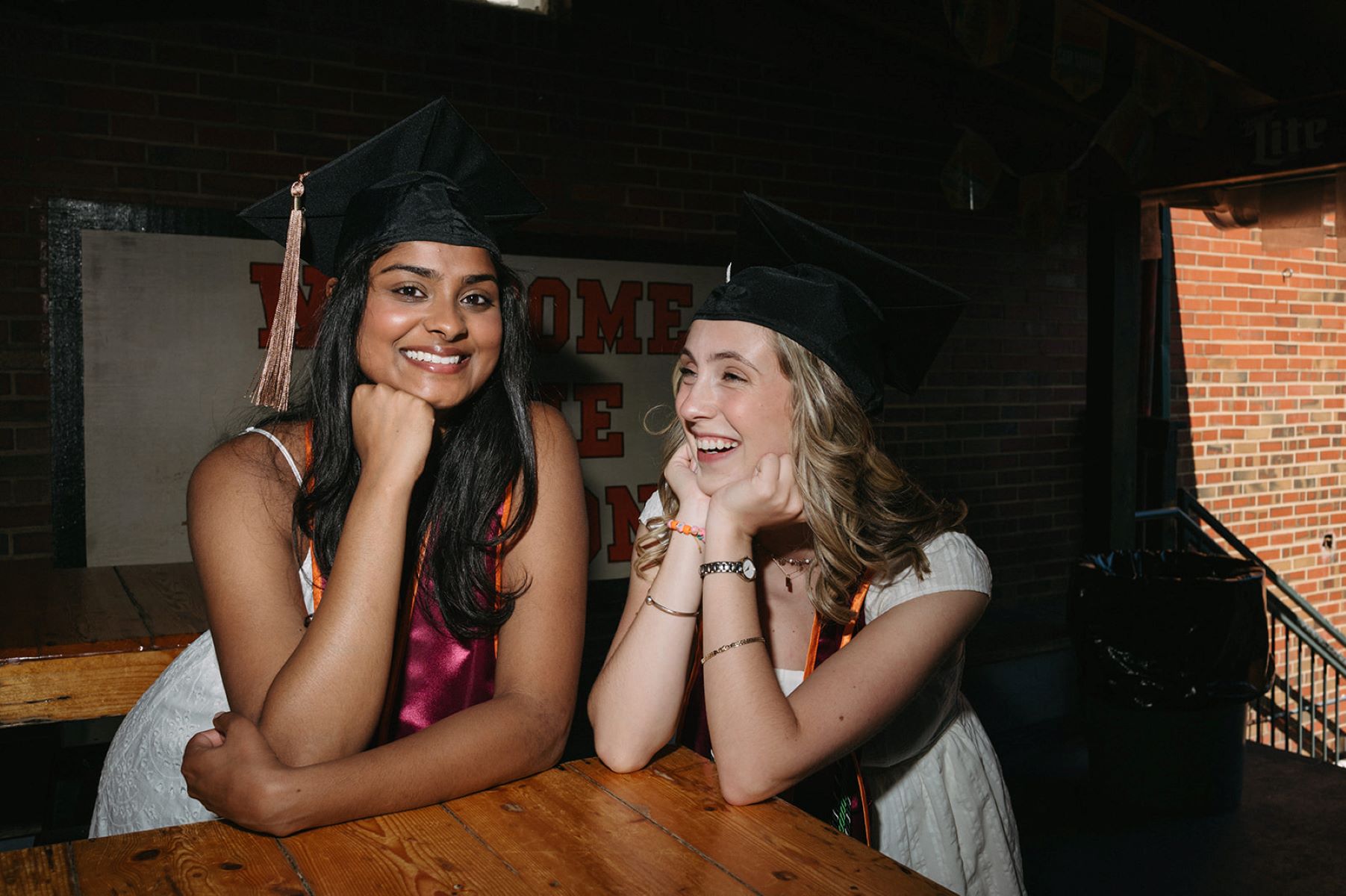 Two girls wearing graduation caps smiling and laughing with each other with their elbows resting on a wooden table 