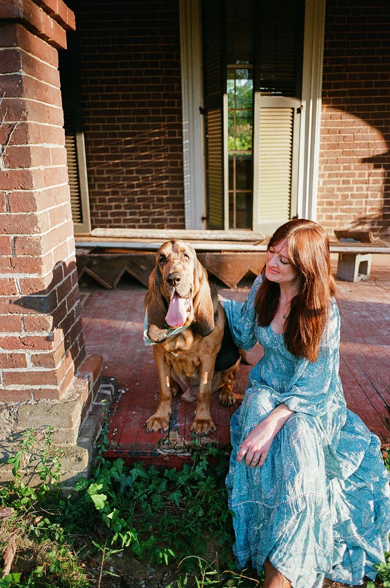 A woman sitting on the deck of a house she is wearing a blue flowy dress and looking at and petting her dog 