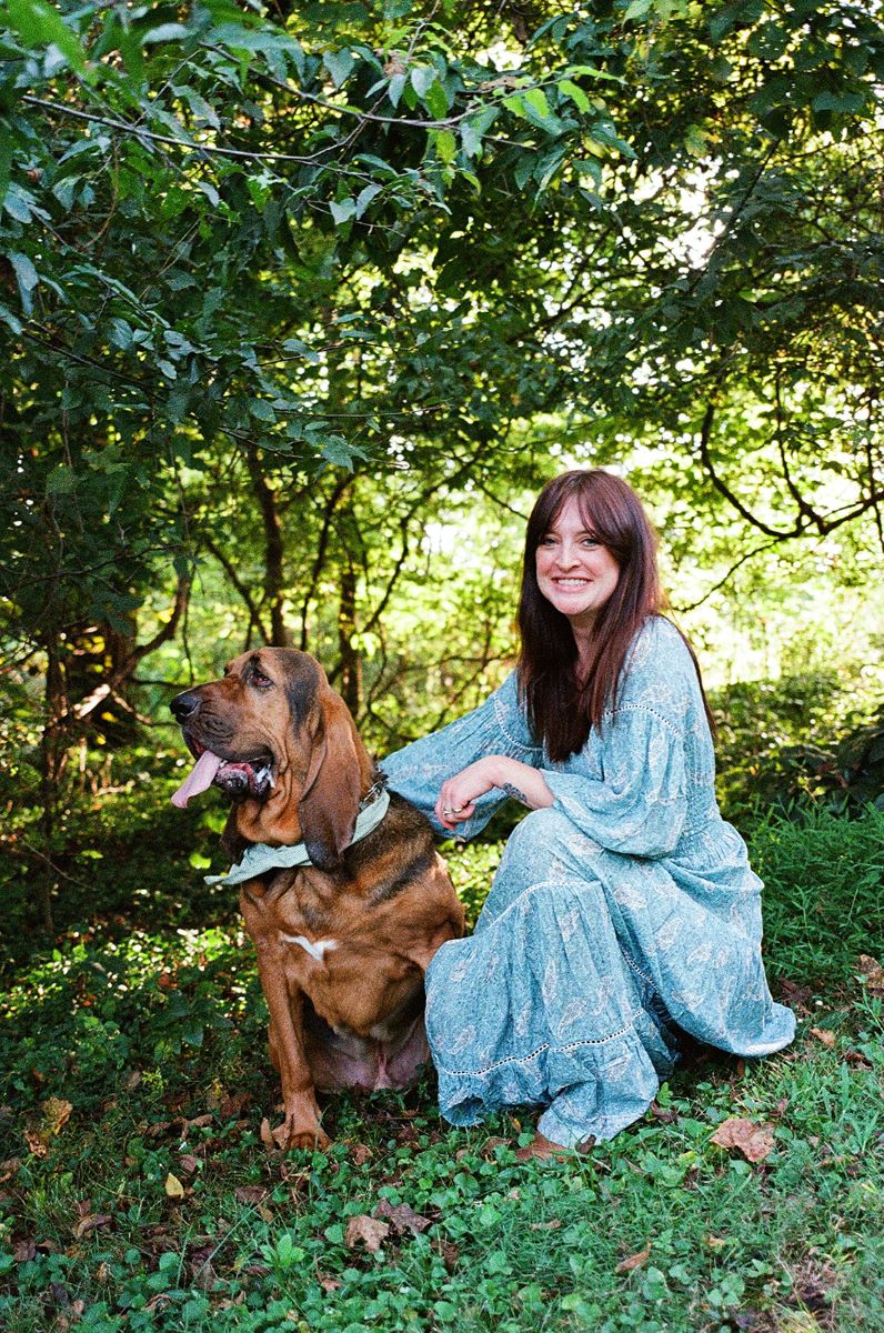 A woman kneeling in the grass smiling and petting their dog 