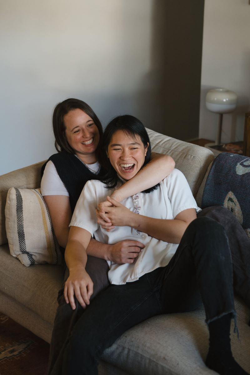 A couple sitting on their couch and laughing one partner is leaning back into the arms of the other and the other partner has their arms wrapped around them