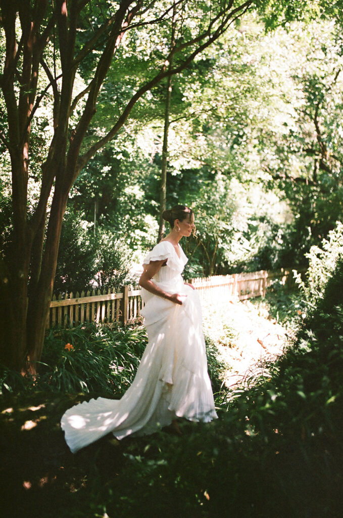 A woman in a wedding dress walks through a heavily forested area with sun shining behind her
