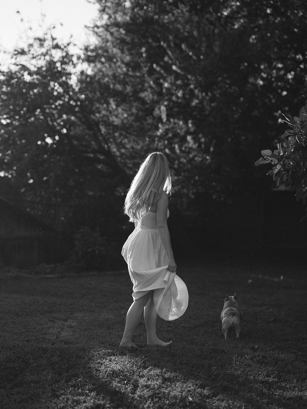 A black and white photo of a woman in a flowy white dress running with her small dog in her backyard